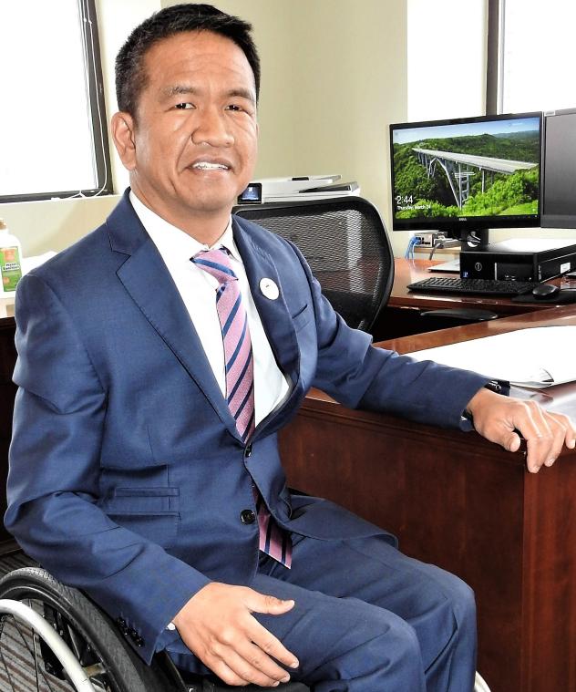 A man in a blue suit with a white shirt and a striped tie. He is using a wheelchair, and is next to a desk.