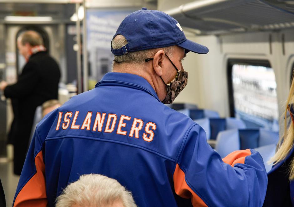 A man in a blue baseball cap and a blue jacket with the word ISLANDERS printed on the back. He is standing on a train.