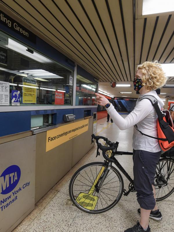 A woman with a bicycle talks with the agent at a subway station booth. She's gesturing toward the turnstiles to her left.
