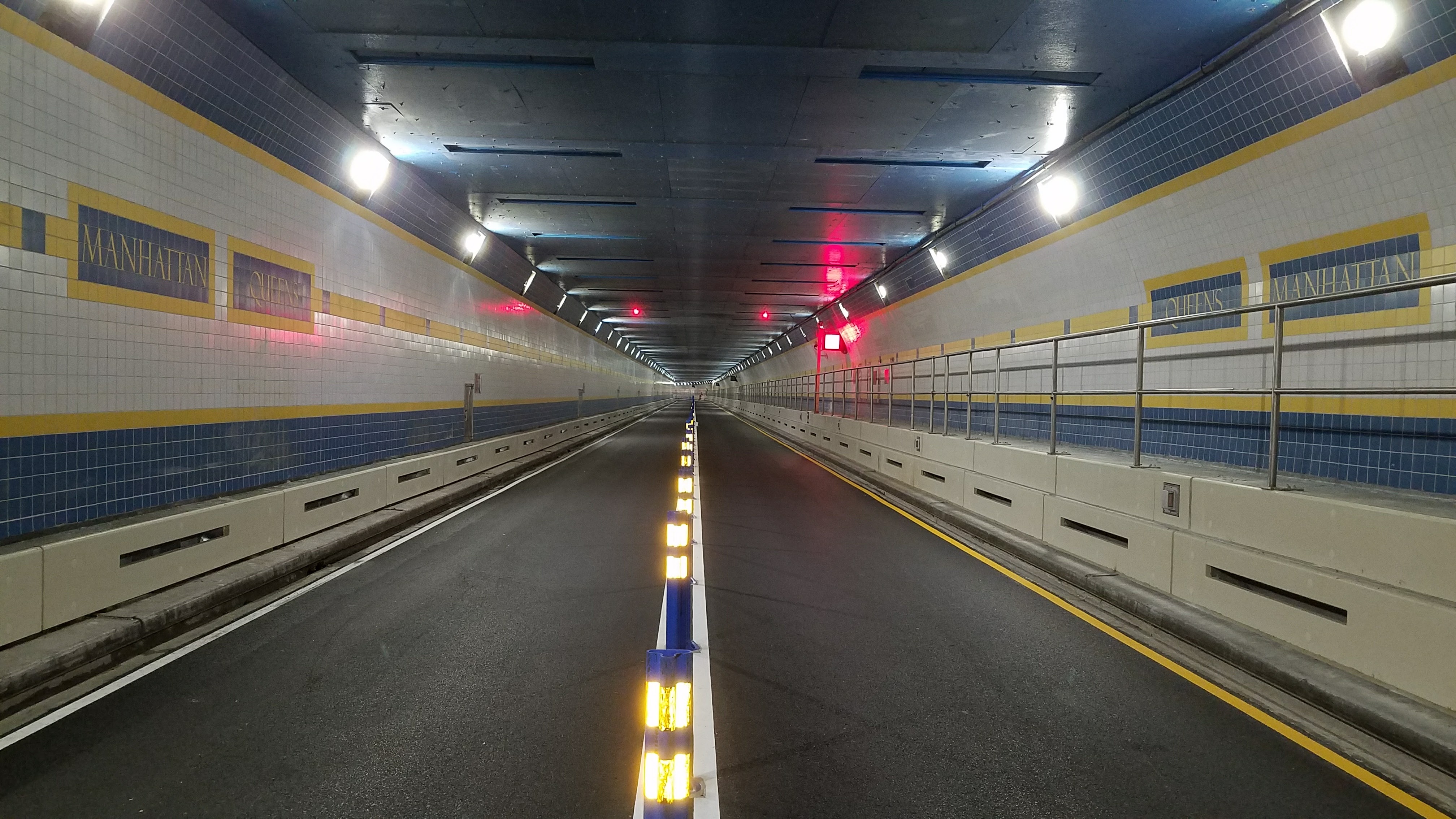 A head-on view of a tunnel designed for vehicular traffic. Blue and yellow subway tiling on the walls says "Manhattan" and "Queens."