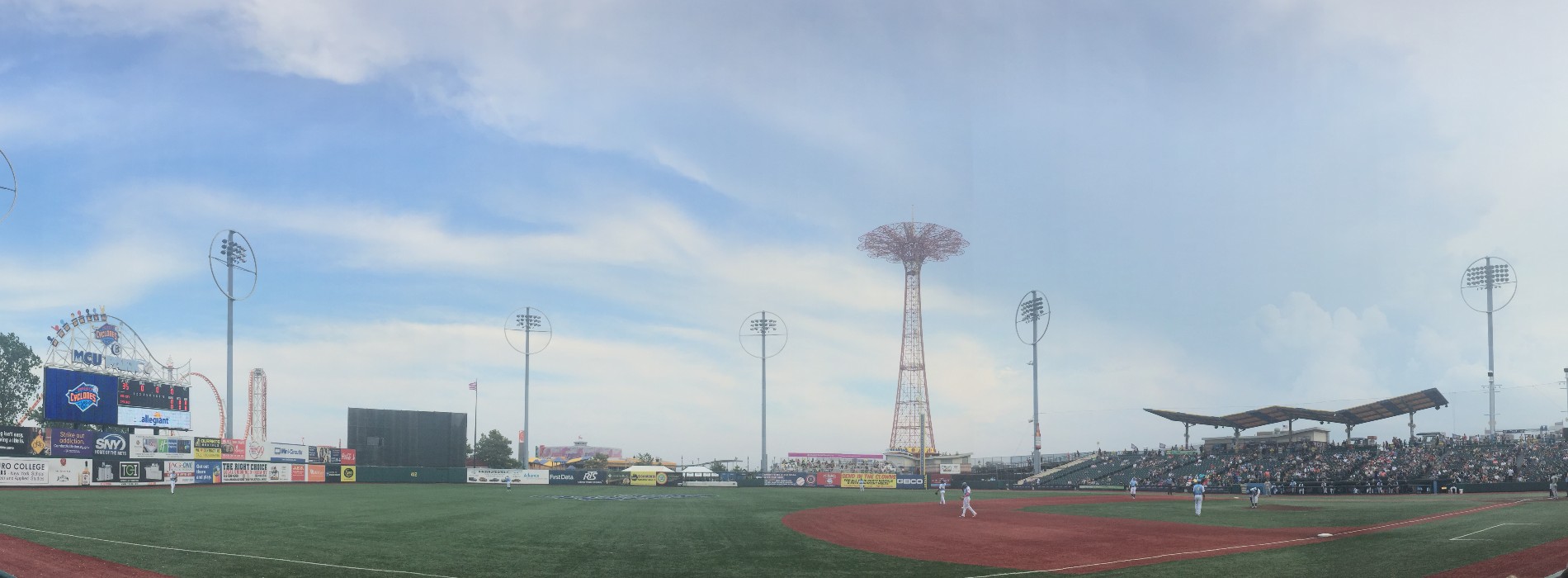 A panoramic view of the Coney Island ballpark under a blue sky, with the Parachute Jump's red tower at the center, beyond right field