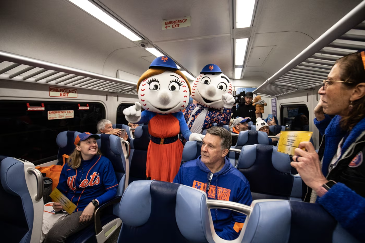Mr. and Mrs. Met stand in the middle of a Long Island Rail Road train car with Mets fans wearing jerseys sitting in the seats