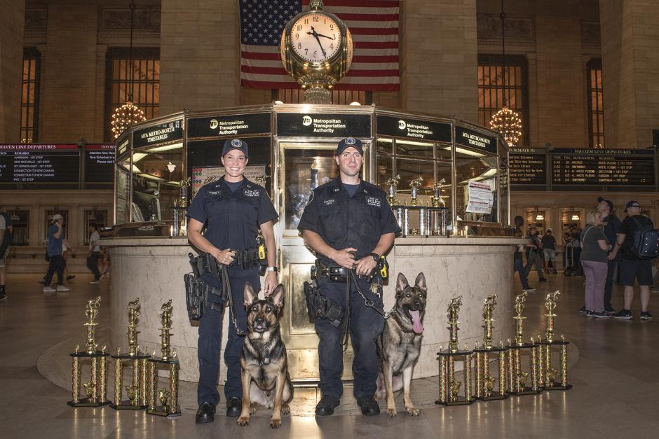 A female and male police officer stand with their K9 partners in front of the information booth at Grand Central Terminal. One of the dogs is sticking its tongue out.